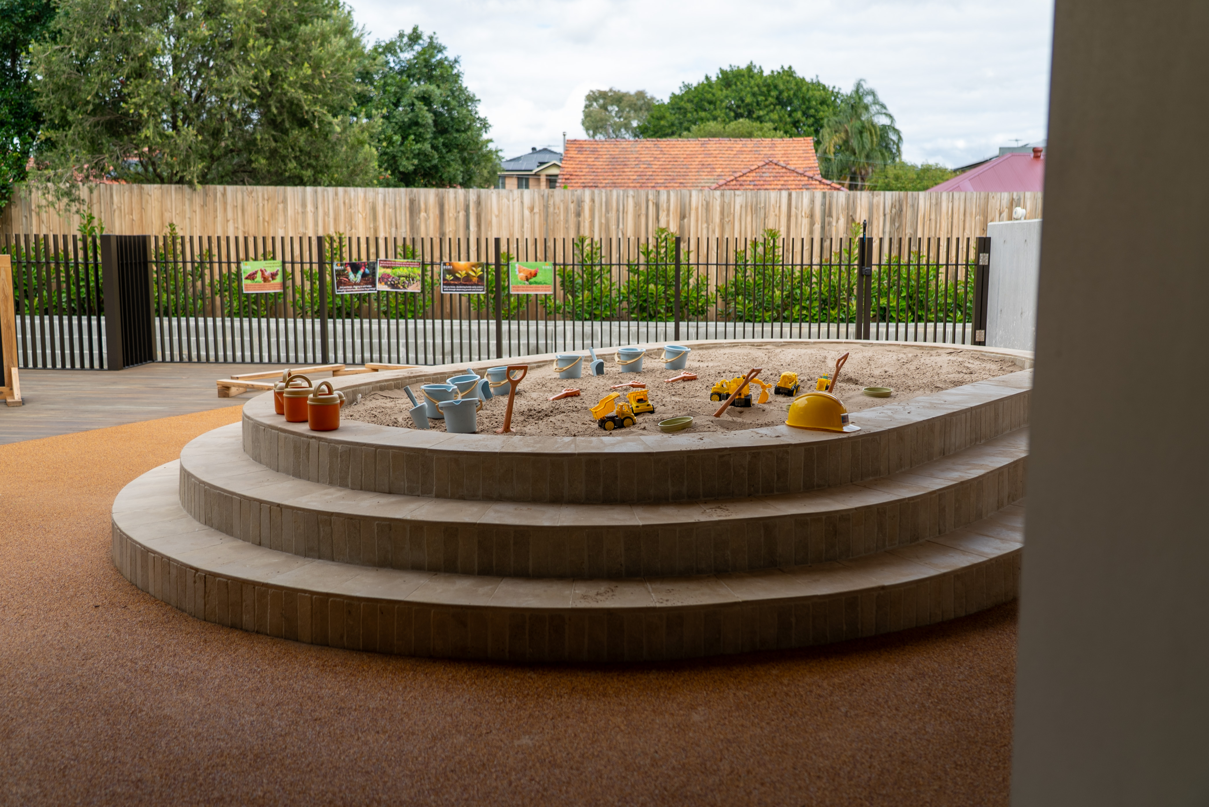 SOMAs circular sandbox with toy trucks, buckets, shovels, and a yellow hard hat around the sand.