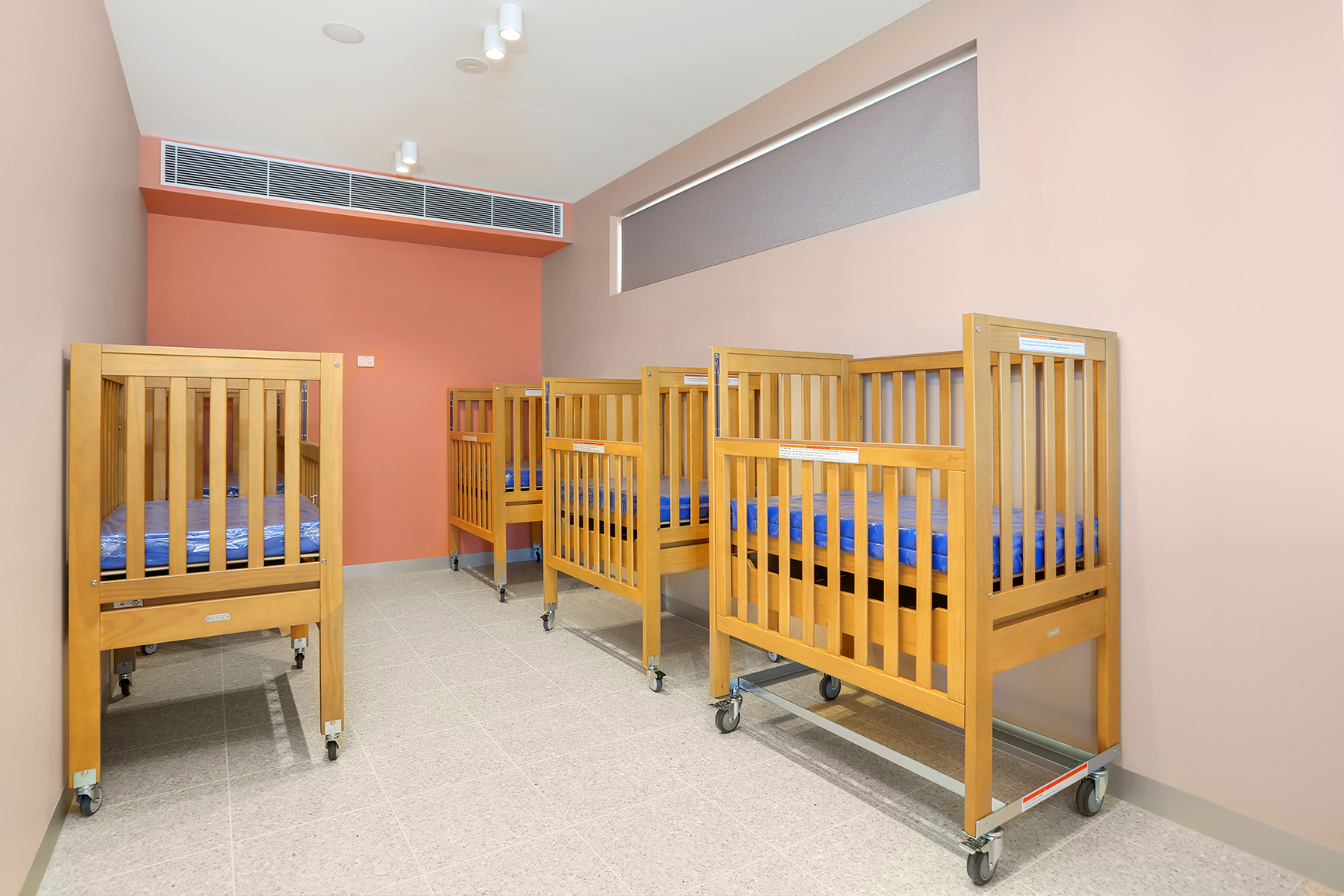 SOMAs nursery room with five wooden baby cots lined up against coral and beige walls.