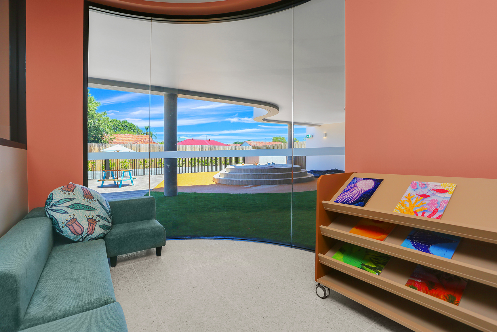 Curved glass wall room with green couch, colorful pillow, and a book cart holding art books.