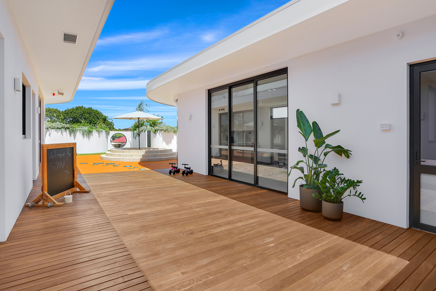 SOMAs modern outdoor play area with wooden deck, play bikes, plants, and shaded steps under blue sky.