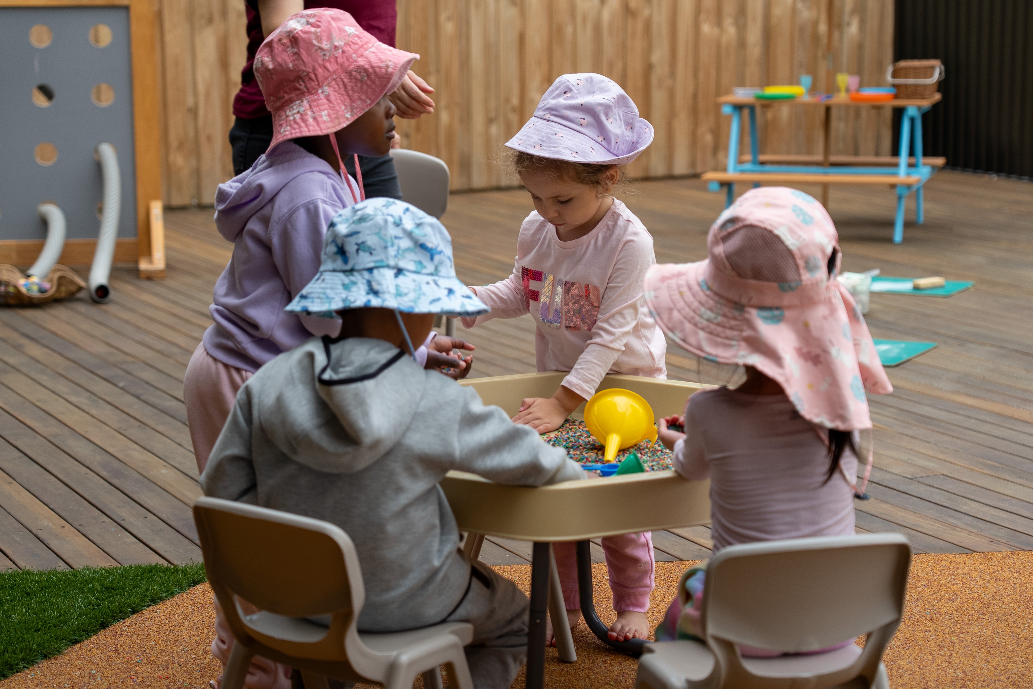 Four young children wearing sun hats playing with sensory beads at a table outdoors.