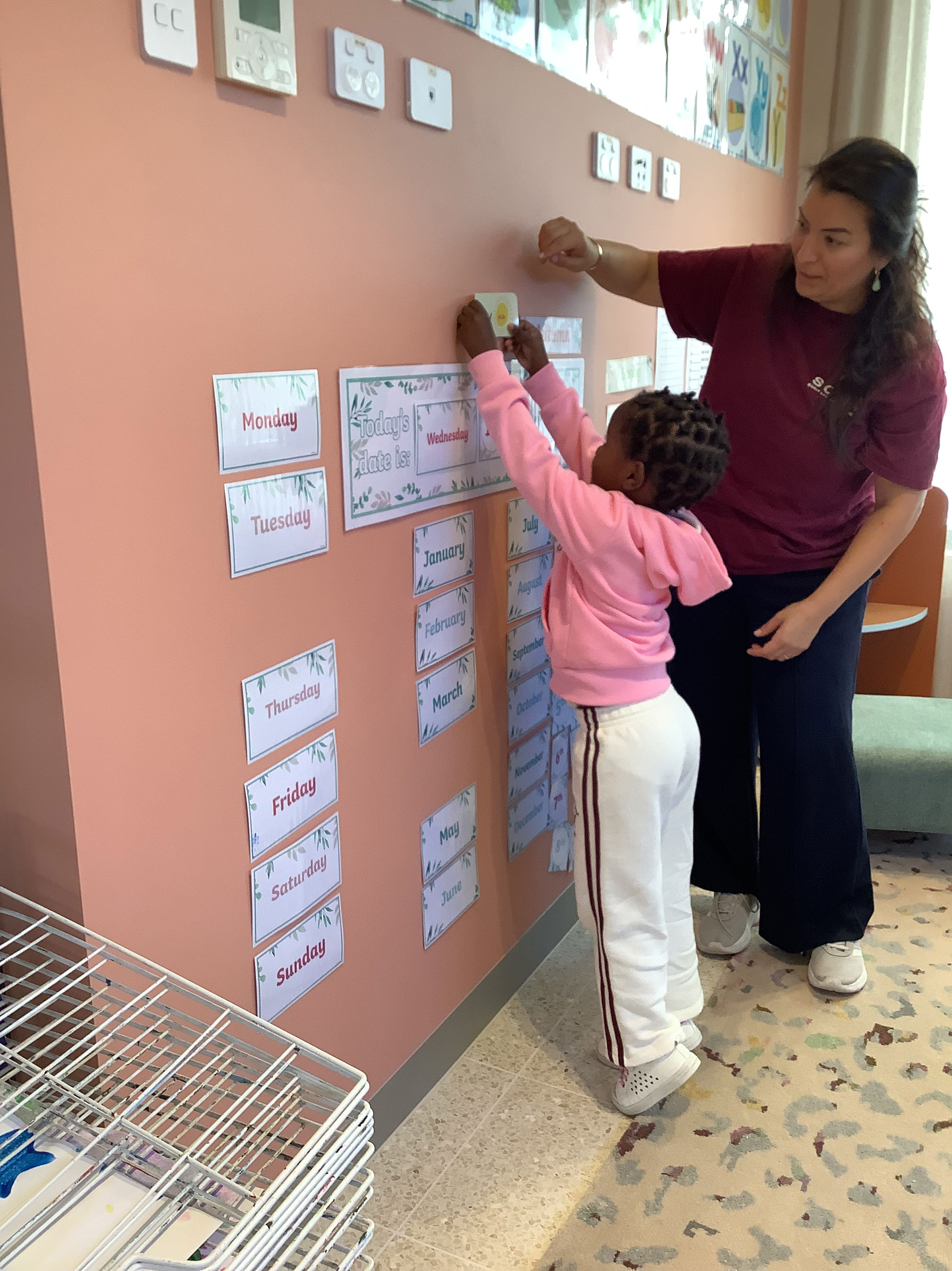Child in pink hoodie placing a card on a classroom wall calendar with a teacher assisting.