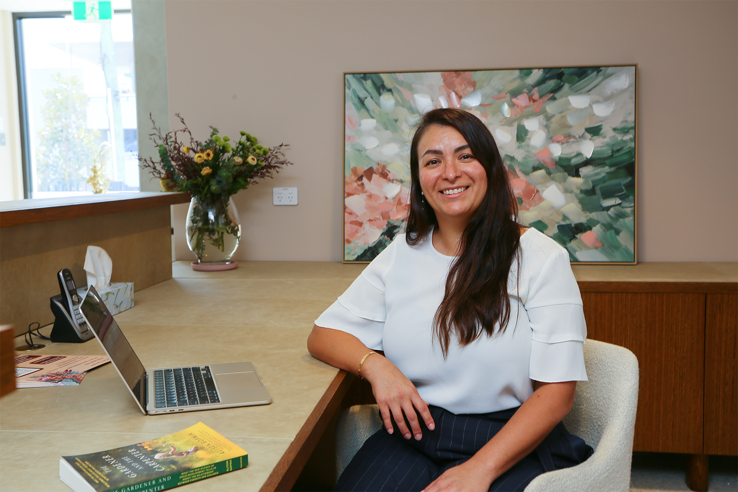 Smiling woman with long hair sitting at desk with laptop and flowers in office setting.