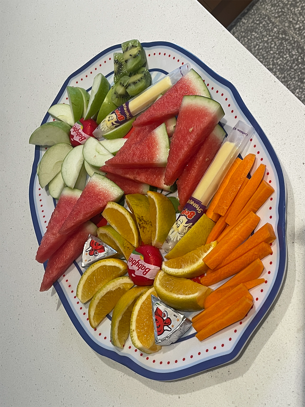 Plate with sliced watermelon, green apple, kiwi, orange, carrot sticks, string cheese, and Babybel cheese.