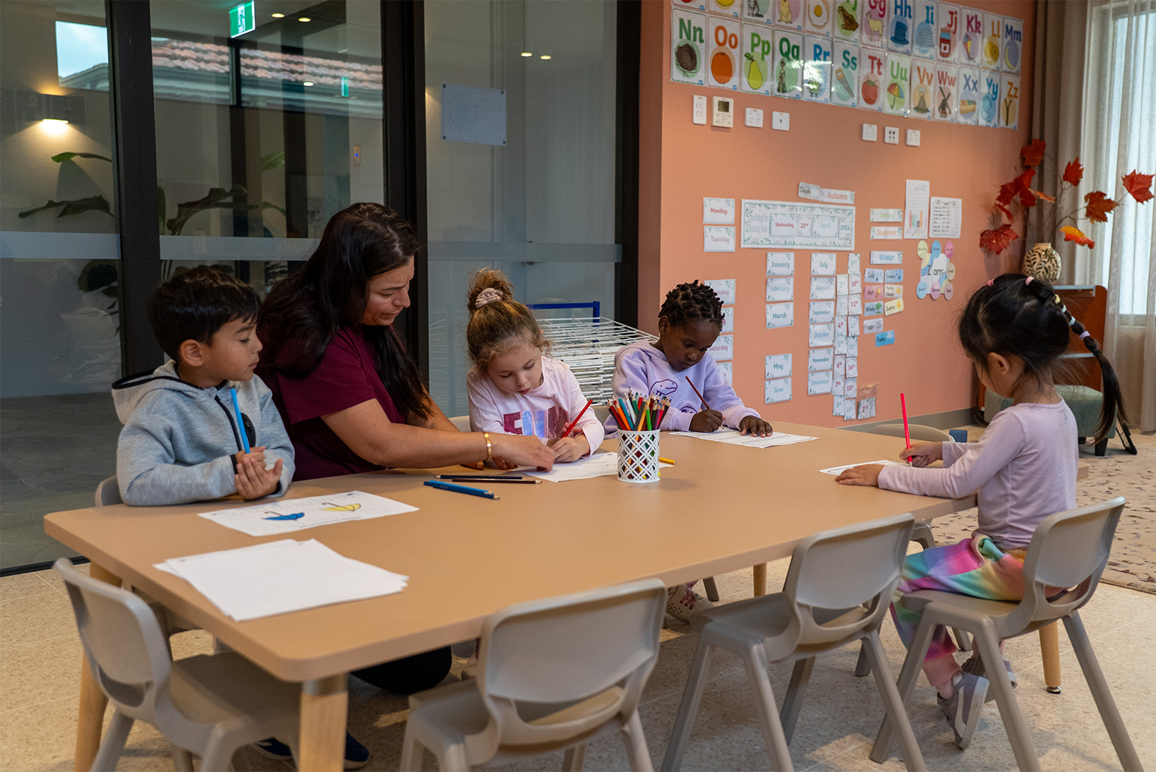 Teacher helps preschool children sitting at a table coloring and drawing in a classroom.