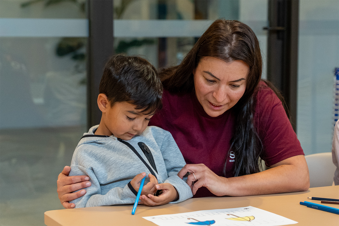 SOMA Director in maroon shirt helps young boy in gray hoodie with a drawing at a table.