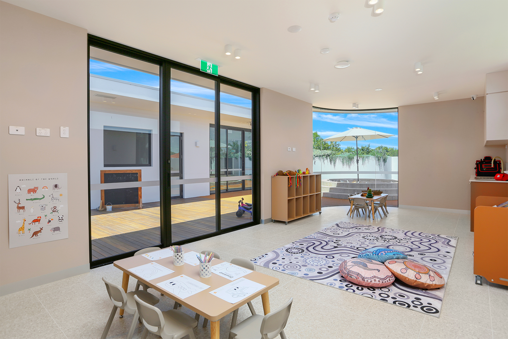 Bright classroom with kids' tables, coloring sheets, cushions on a patterned rug, and large windows to outdoors.