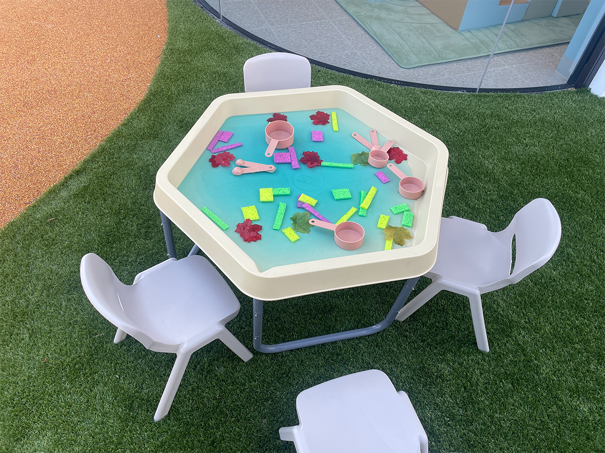 Hexagonal water play table with plastic cups, spoons, colorful sponges, and leaves surrounded by four plastic chairs.