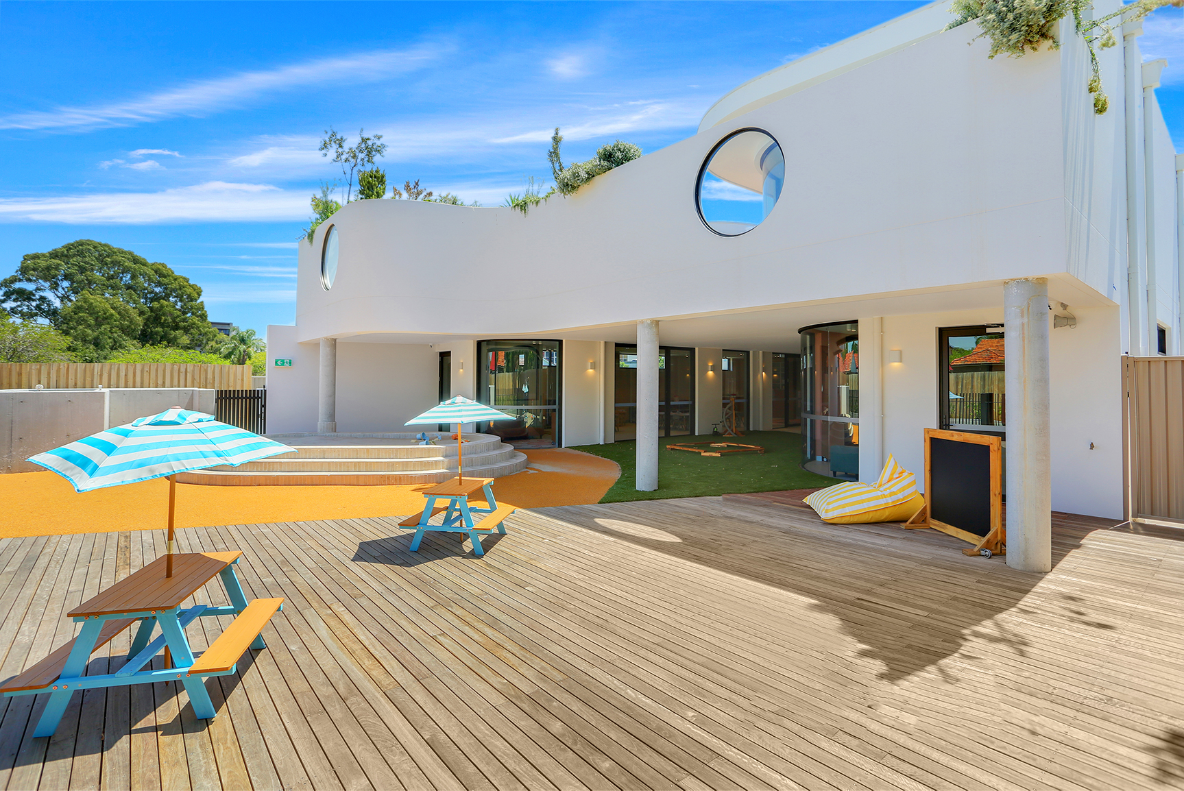 Modern outdoor patio with two blue-striped umbrellas over picnic tables and a white building with round windows.