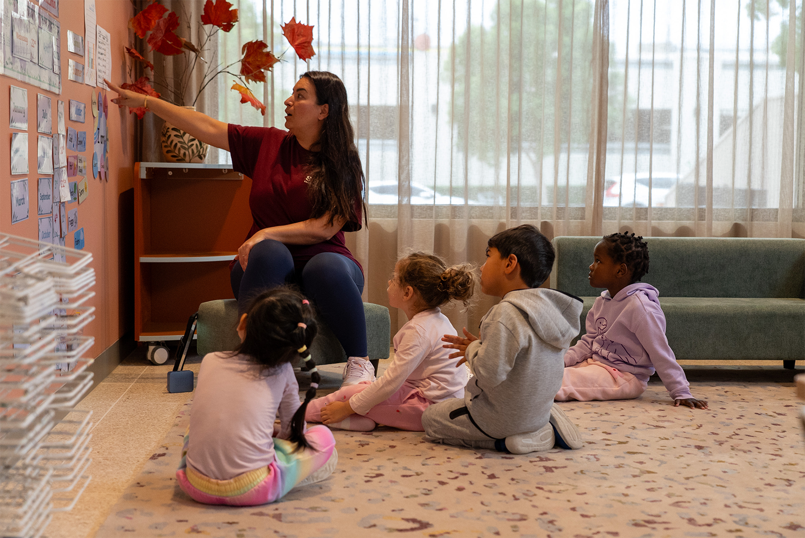 Teacher sitting on a stool pointing at classroom wall while four children sit on the floor watching.