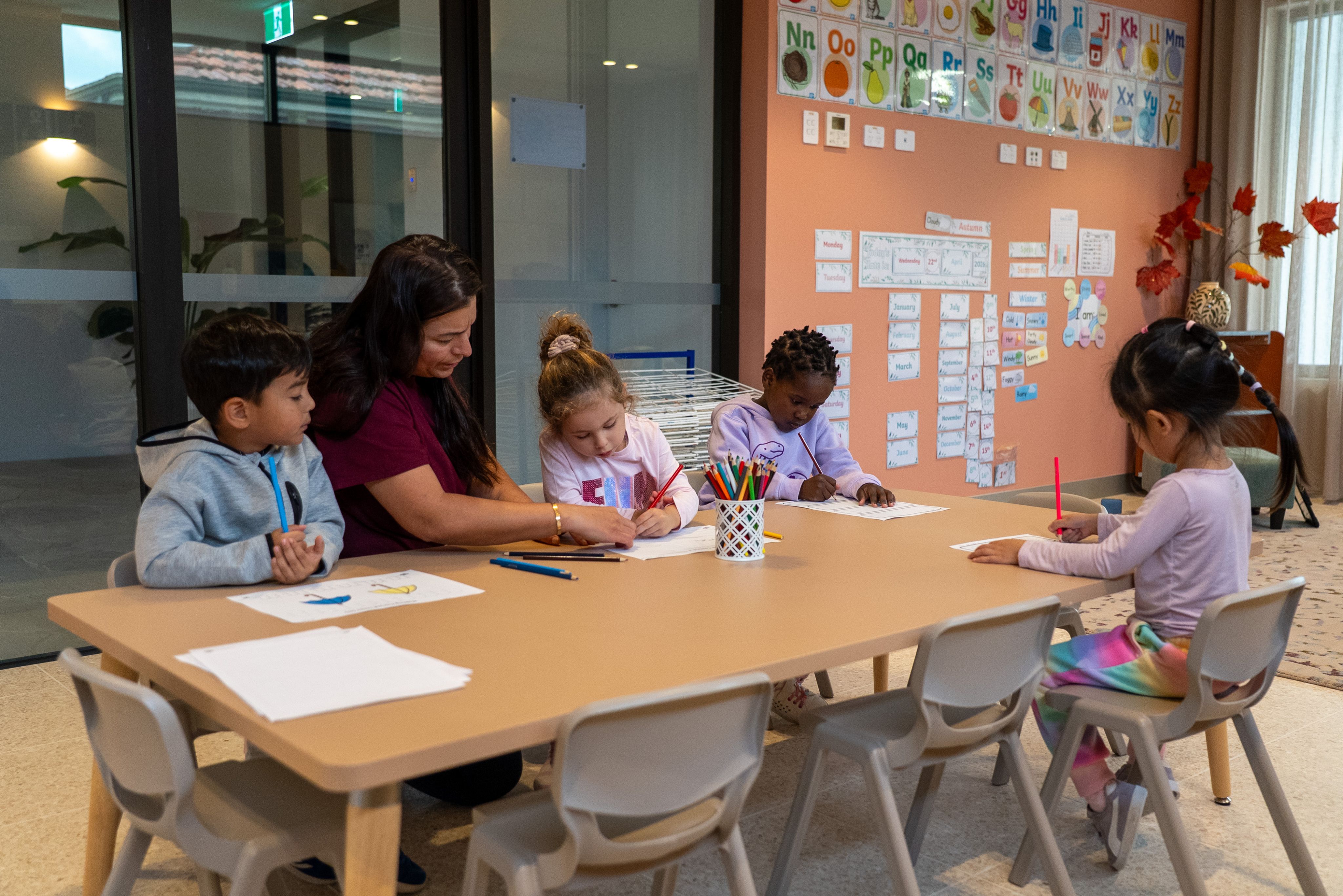 Teacher helping young children coloring at a classroom table with alphabet posters on the wall.