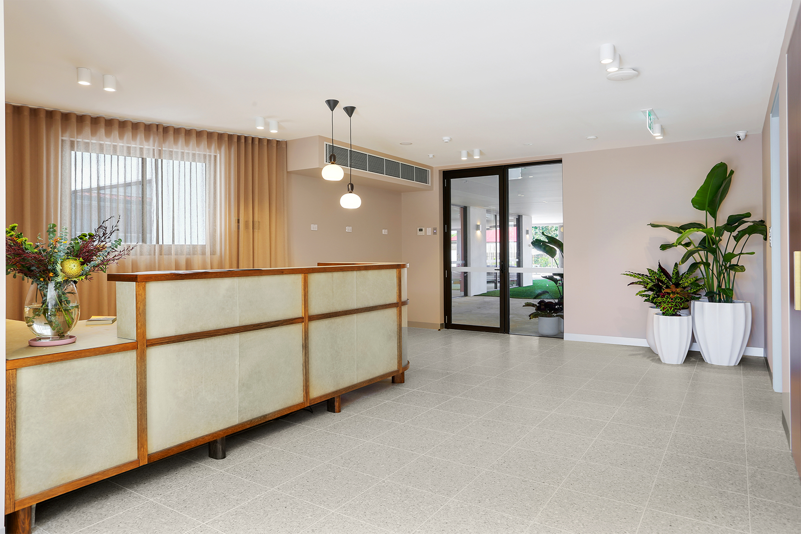 Reception area with wooden counter, pendant lights, large window with sheer curtains, and potted plants by glass doors.
