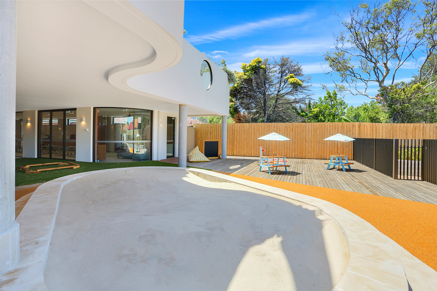 Outdoor play area with empty sandpit, two picnic tables with umbrellas, wooden fence, and greenery behind.