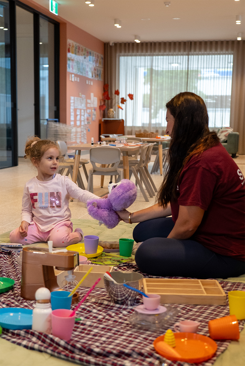 A woman and a young girl play with a purple stuffed toy in a room with toys and tables.
