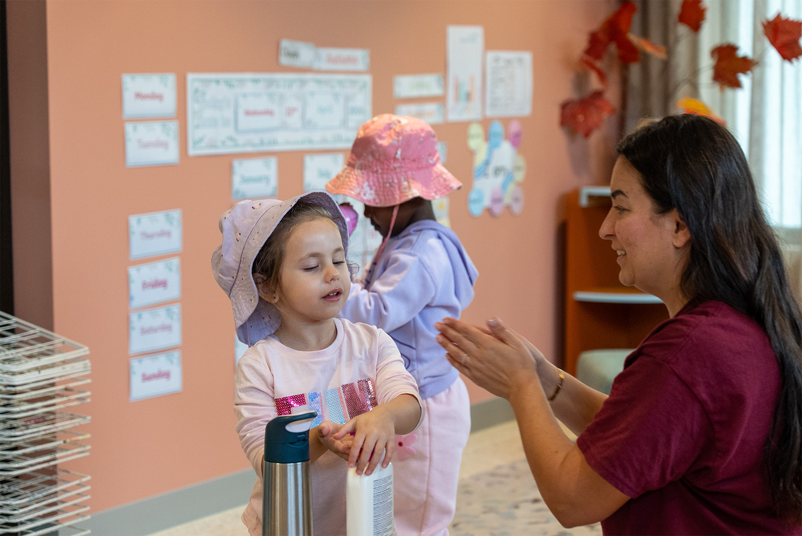 Teacher clapping hands while young girls in sun hats apply lotion indoors with educational posters behind.