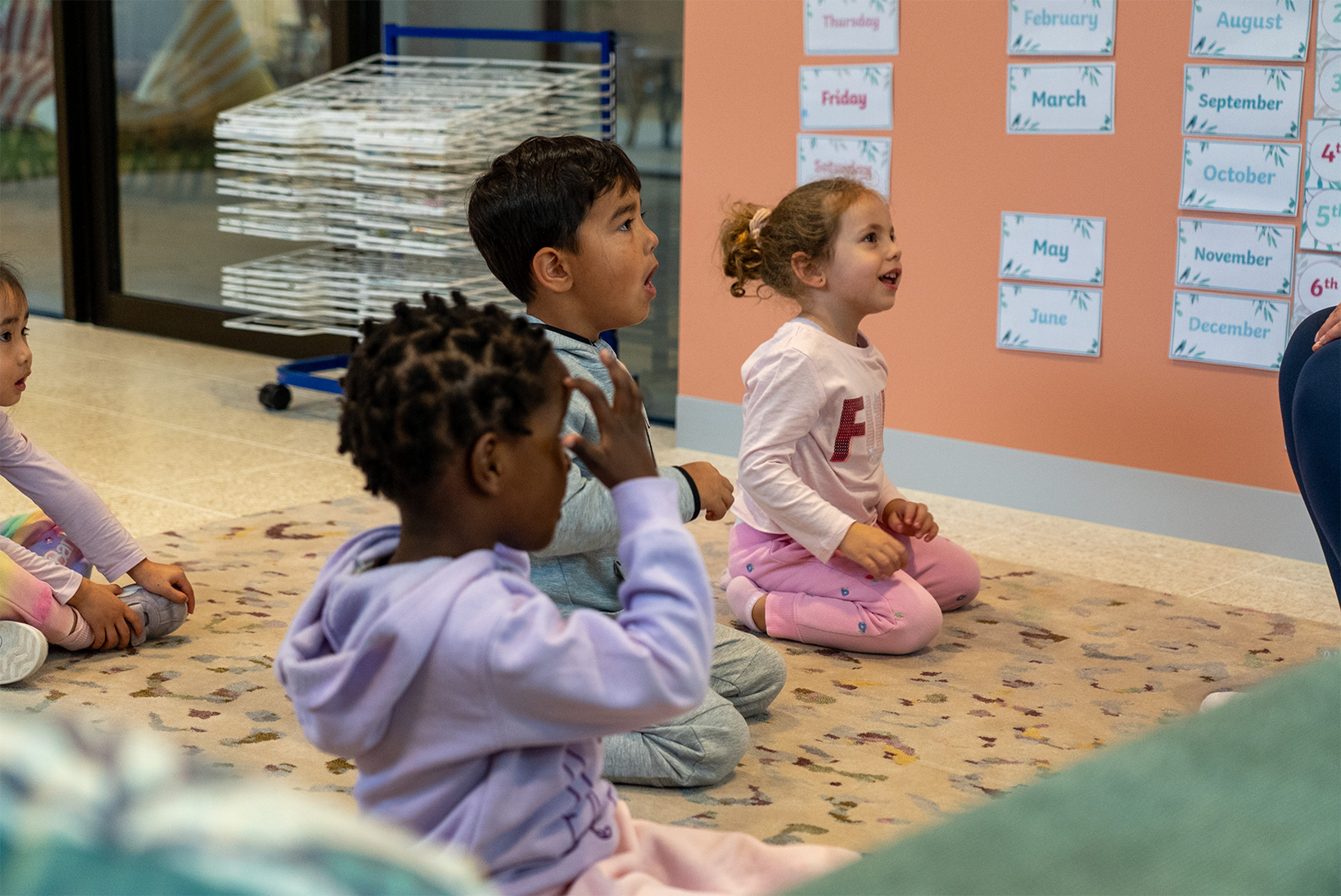 Three children sitting on a rug in a classroom, attentively looking forward at a lesson.