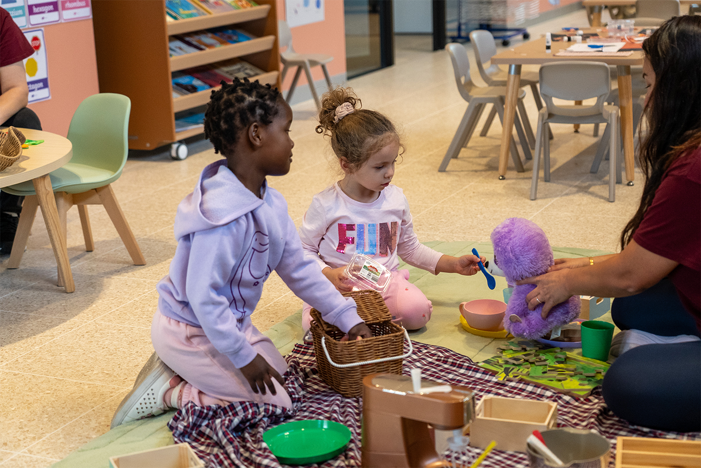 Two young girls play with toy kitchen and stuffed animal while an adult assists them indoors.