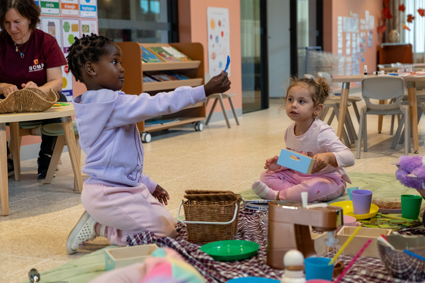Two young children playing with toy kitchen and blocks on a mat in a bright classroom.