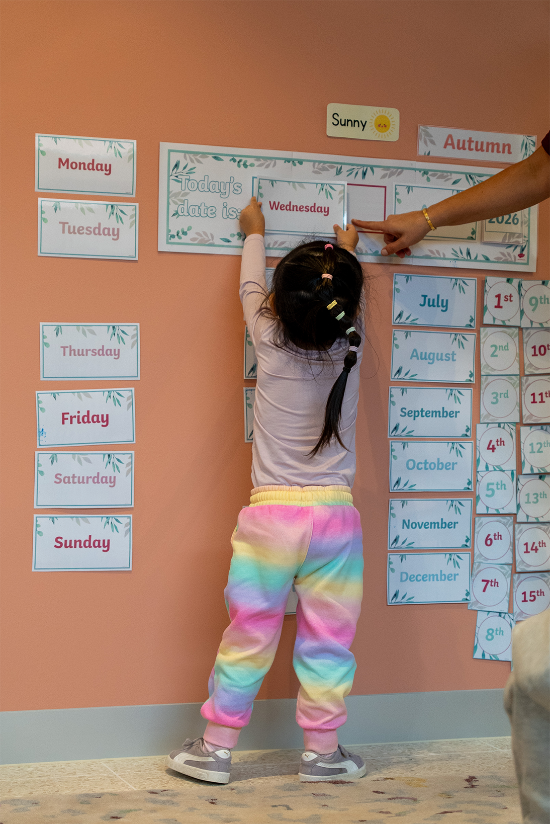 Child in rainbow pants placing a 'Wednesday' card on a classroom calendar with an adult pointing.