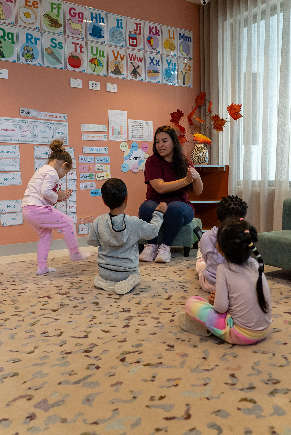 Teacher clapping hands with four young children sitting and standing on a carpet in a colorful classroom.