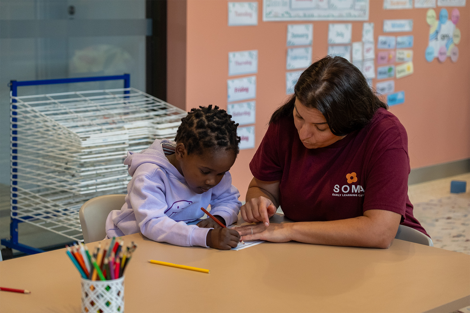 Teacher in maroon shirt guiding a child in a purple hoodie with drawing at a table in classroom.