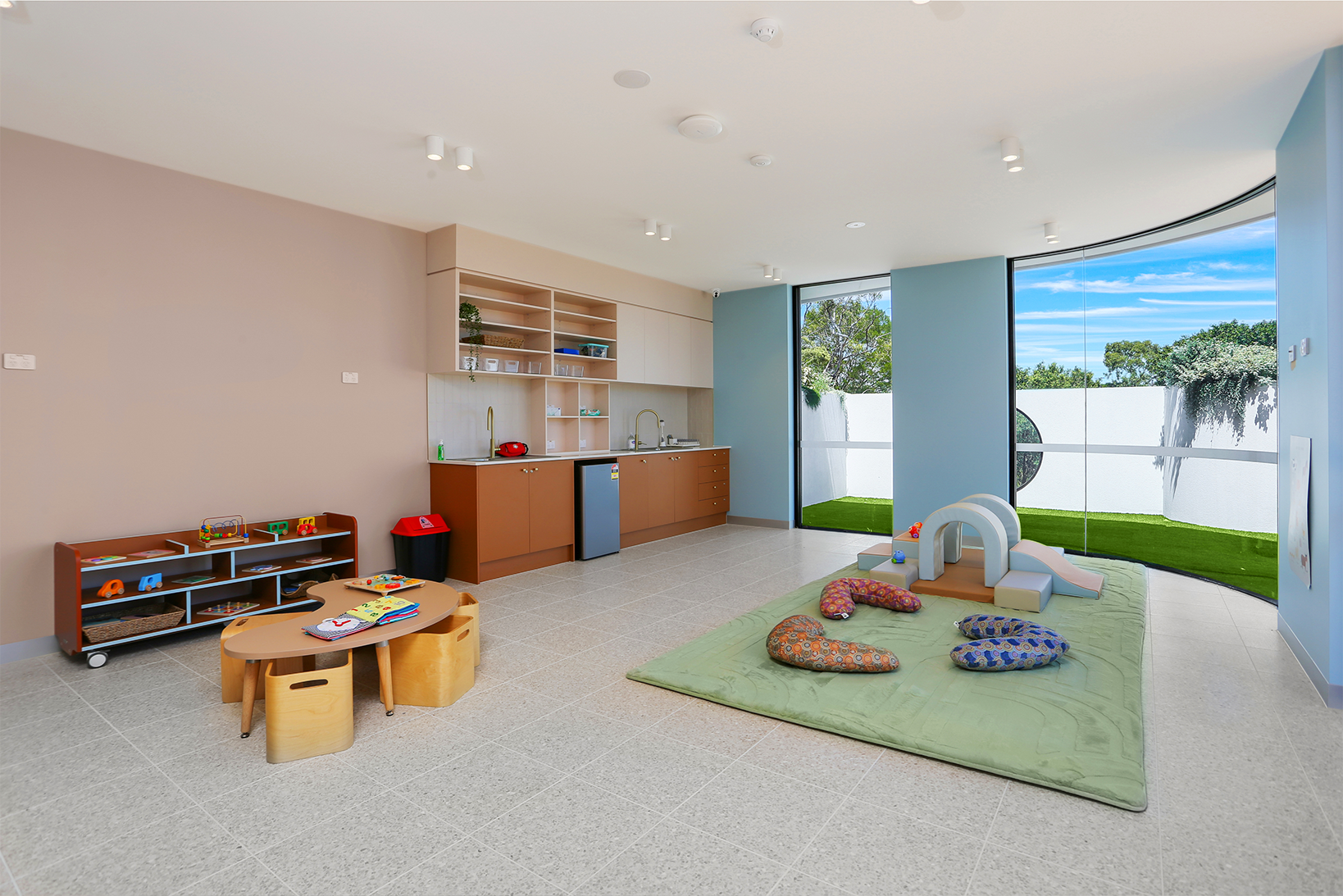 Bright playroom with toys, low tables, cushioned seating, kitchenette, and large windows overlooking grass yard.
