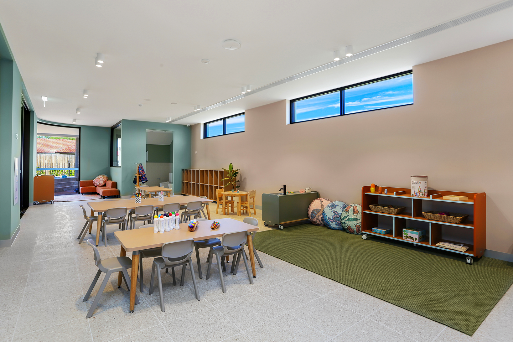 Bright classroom with child-sized tables, chairs, shelves, cushions, and a green carpet area under windows.