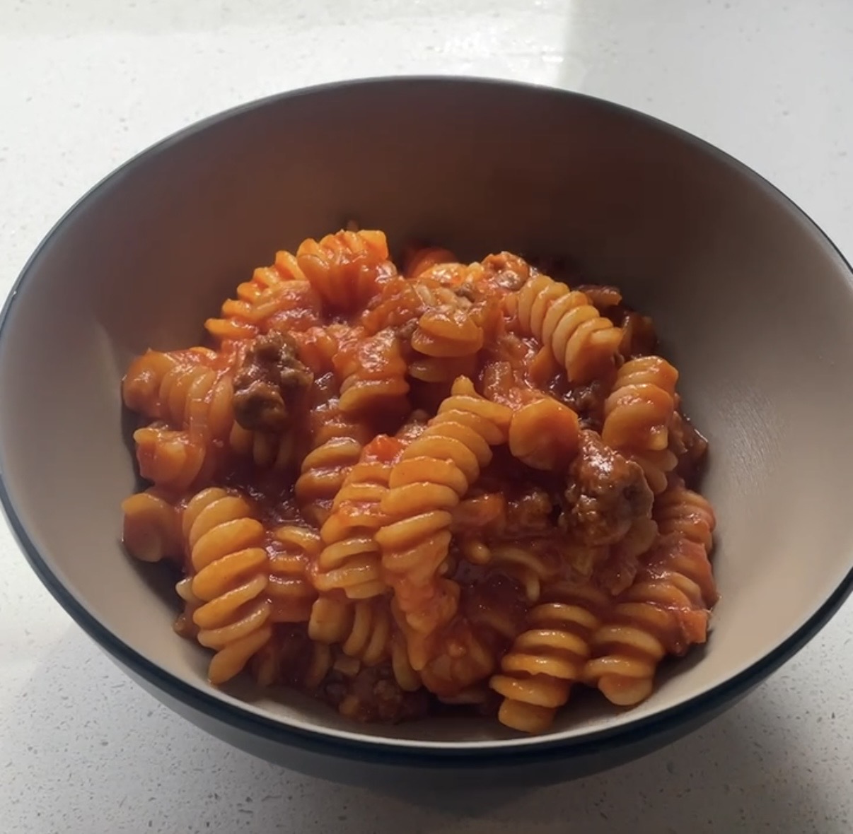 Bowl of rotini pasta mixed with meat and tomato sauce on a white countertop.