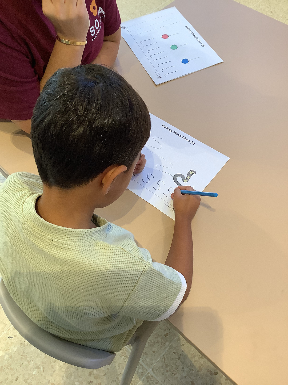 Child in light green shirt tracing wavy lines on a worksheet while an adult watches nearby.