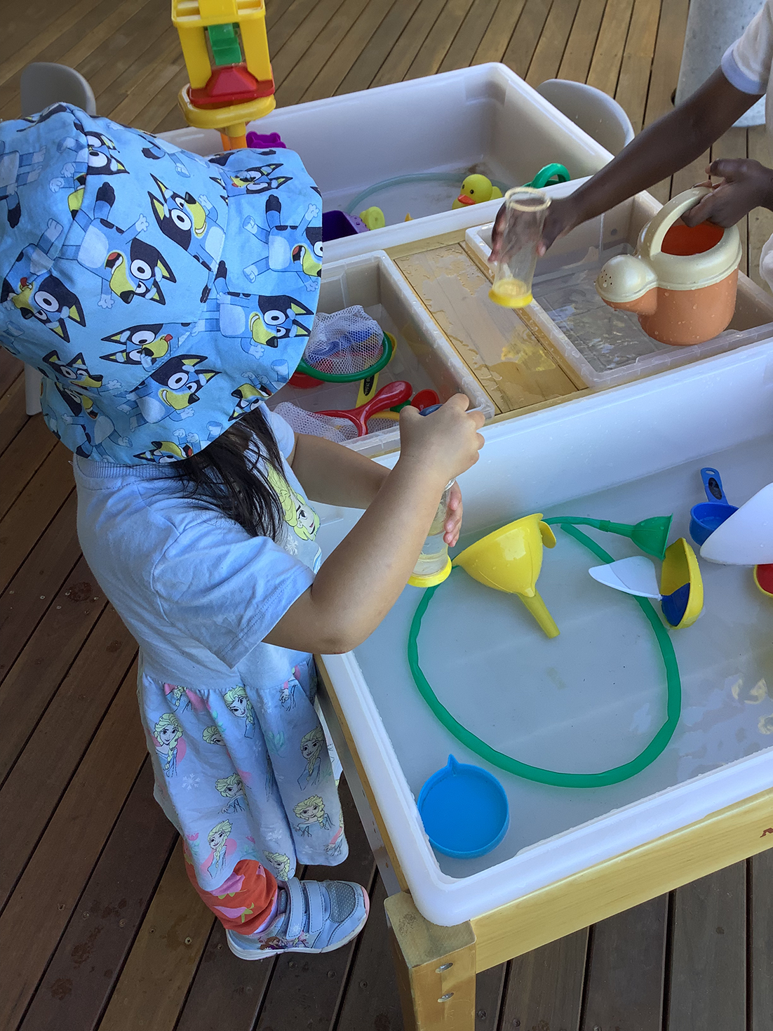 Two children play with water table toys, one wearing a blue cartoon hat and dress.