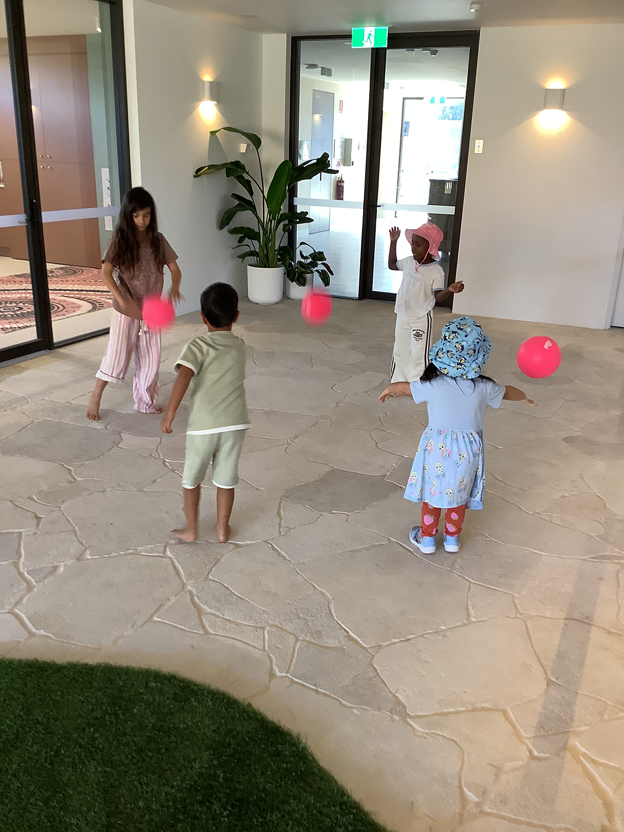 Four children playing with pink balls in a spacious indoor area with glass doors and plants.