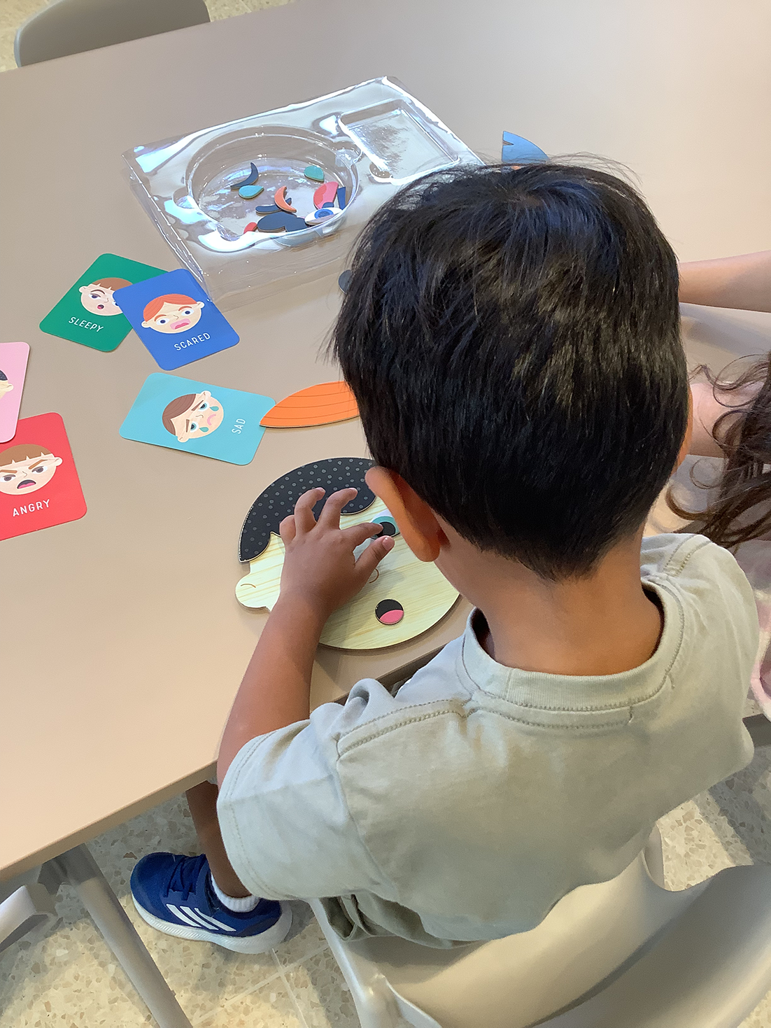 Child assembling a face puzzle with emotion cards labeled sleepy, scared, sad, and angry on the table.