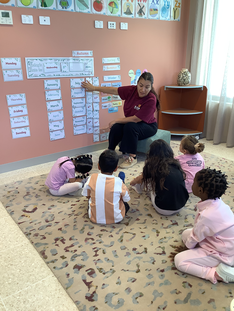 Teacher sitting on a stool pointing to calendar cards on the wall as young children sit on a rug listening.