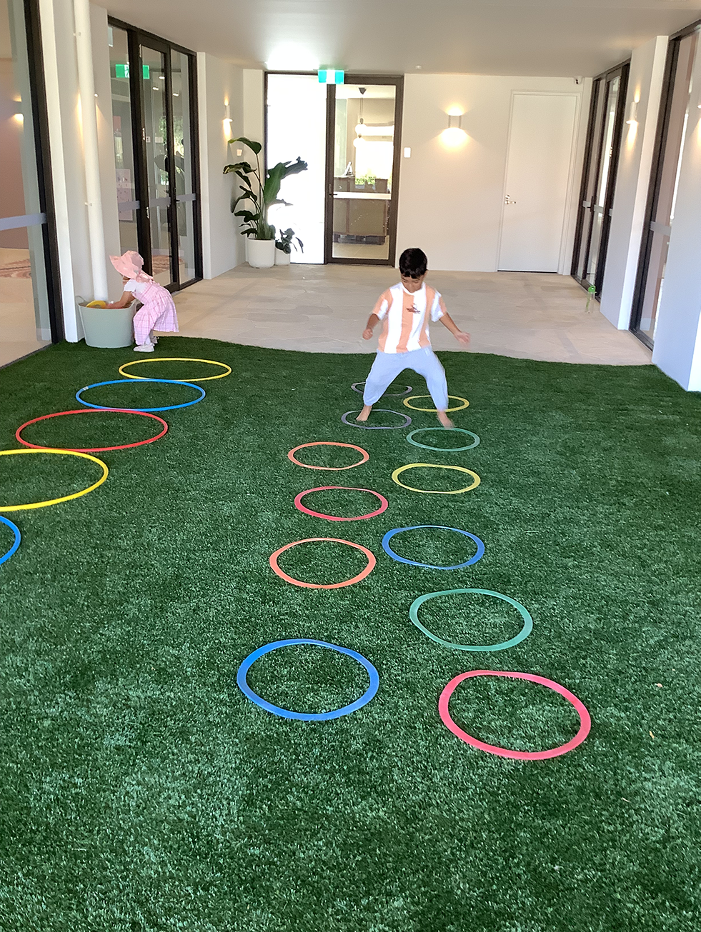 Two children playing indoors on green turf with colorful plastic rings arranged on the ground.