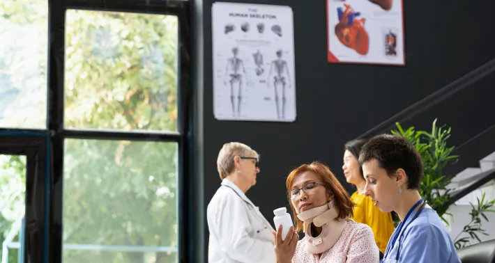 A female patient wearing a neck brace is consulting with a doctor holding a medication bottle in a medical office with another patient and doctor in the background.