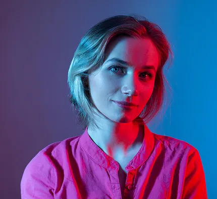 Young woman with short hair wearing a pink shirt illuminated by blue and red lighting against a dark background.