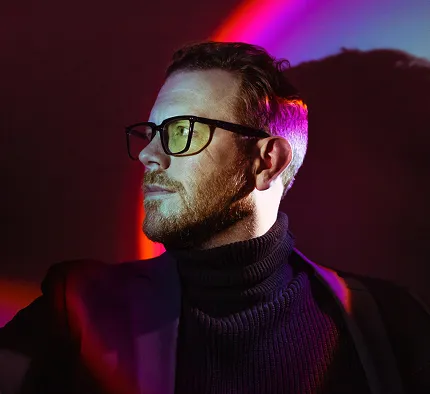 Man with glasses and beard wearing a black turtleneck and jacket, illuminated by colorful red and purple lighting with shadow on the wall.