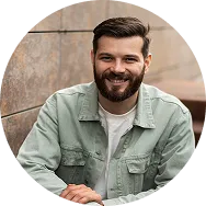 Smiling man with beard wearing a light green button-up shirt sitting outdoors.