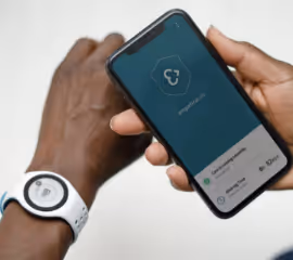 White Empatica EmbracePlus on a person's wrist on the left and someone holding a phone with a health app open on the phone.