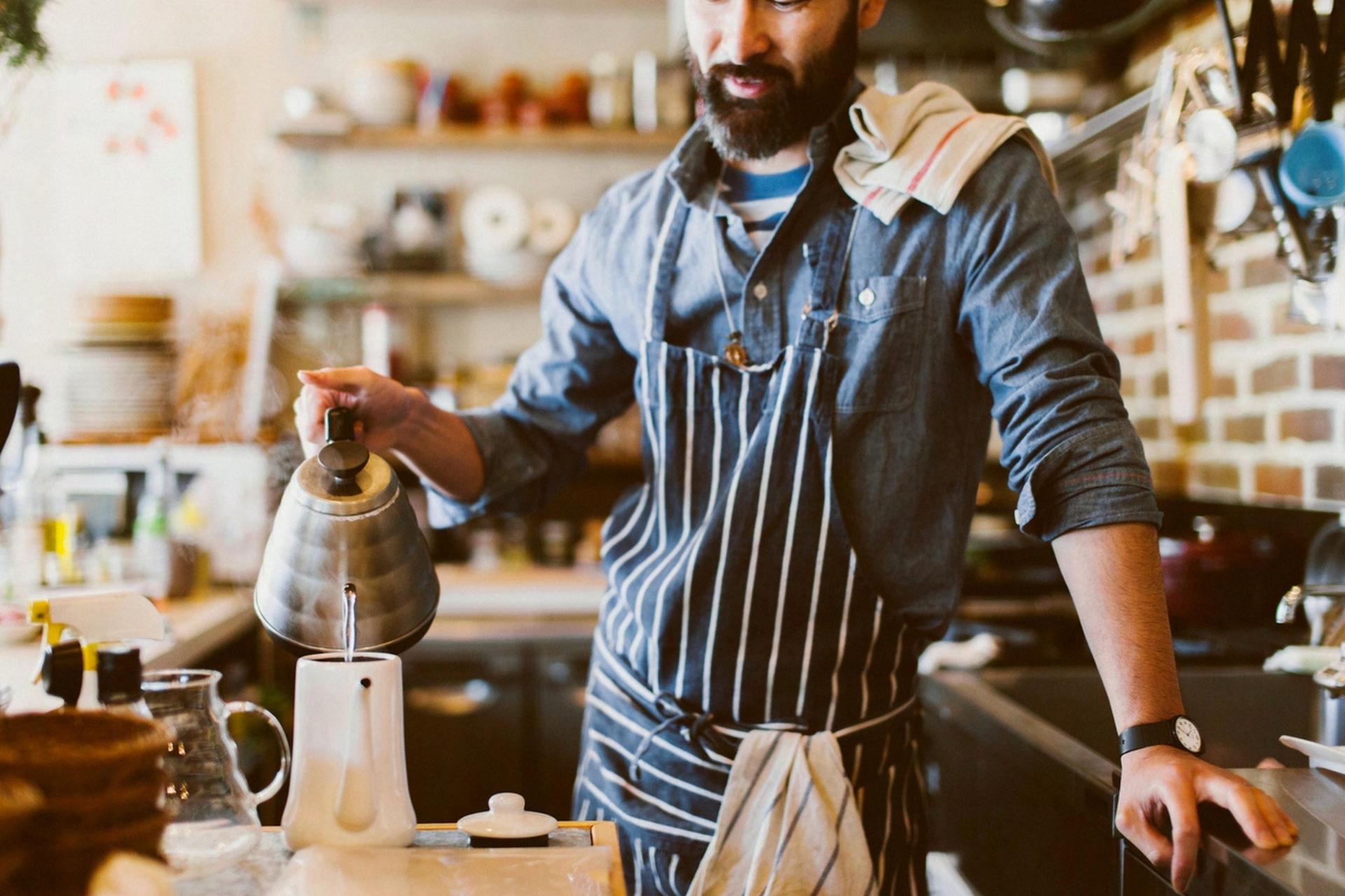 Man in striped apron pouring hot water from kettle into white coffee dripper in kitchen.