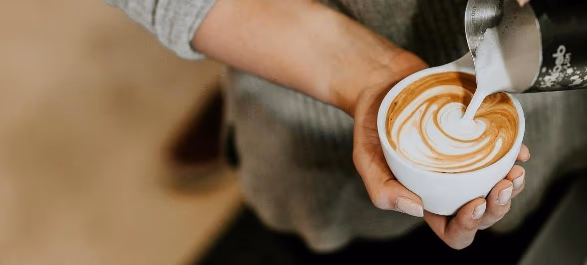 Person pouring milk into a cup of cappuccino with latte art swirl.