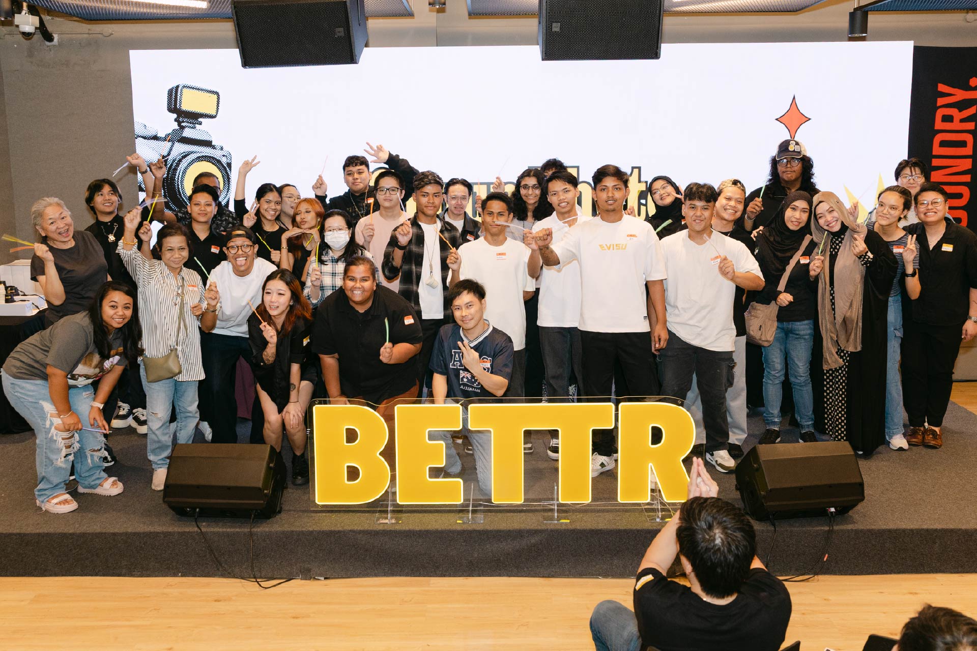 Group of diverse people smiling and posing behind large yellow letters spelling BETTR on stage.