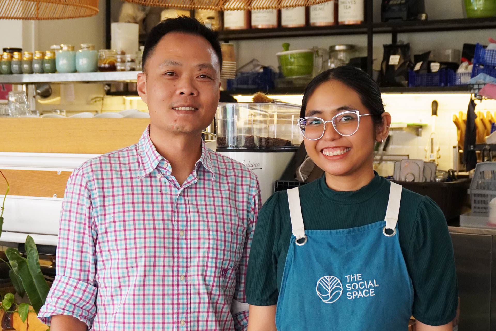 Smiling man in checkered shirt and woman in glasses wearing The Social Space apron in a cafe.