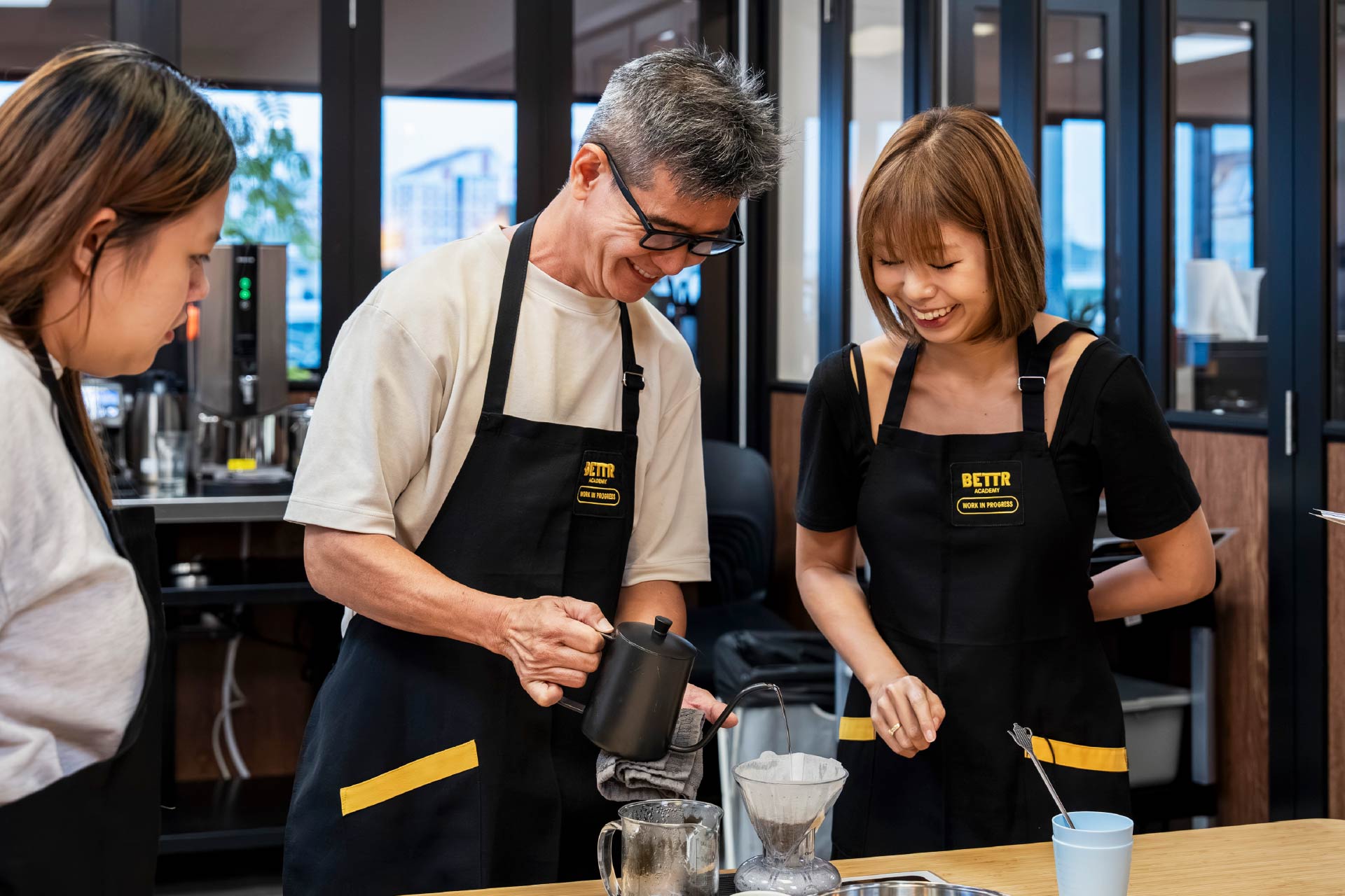 Three people wearing black aprons preparing coffee with a pour-over coffee maker at a workshop.