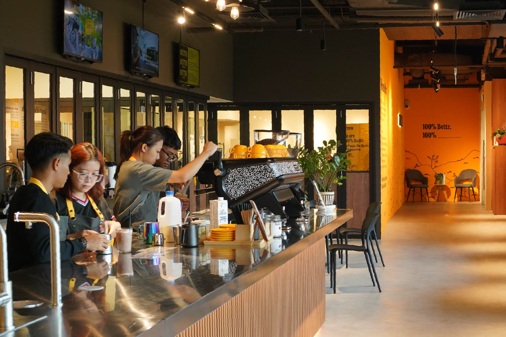 Four baristas preparing drinks behind a modern coffee bar with yellow cups and bags of coffee beans.