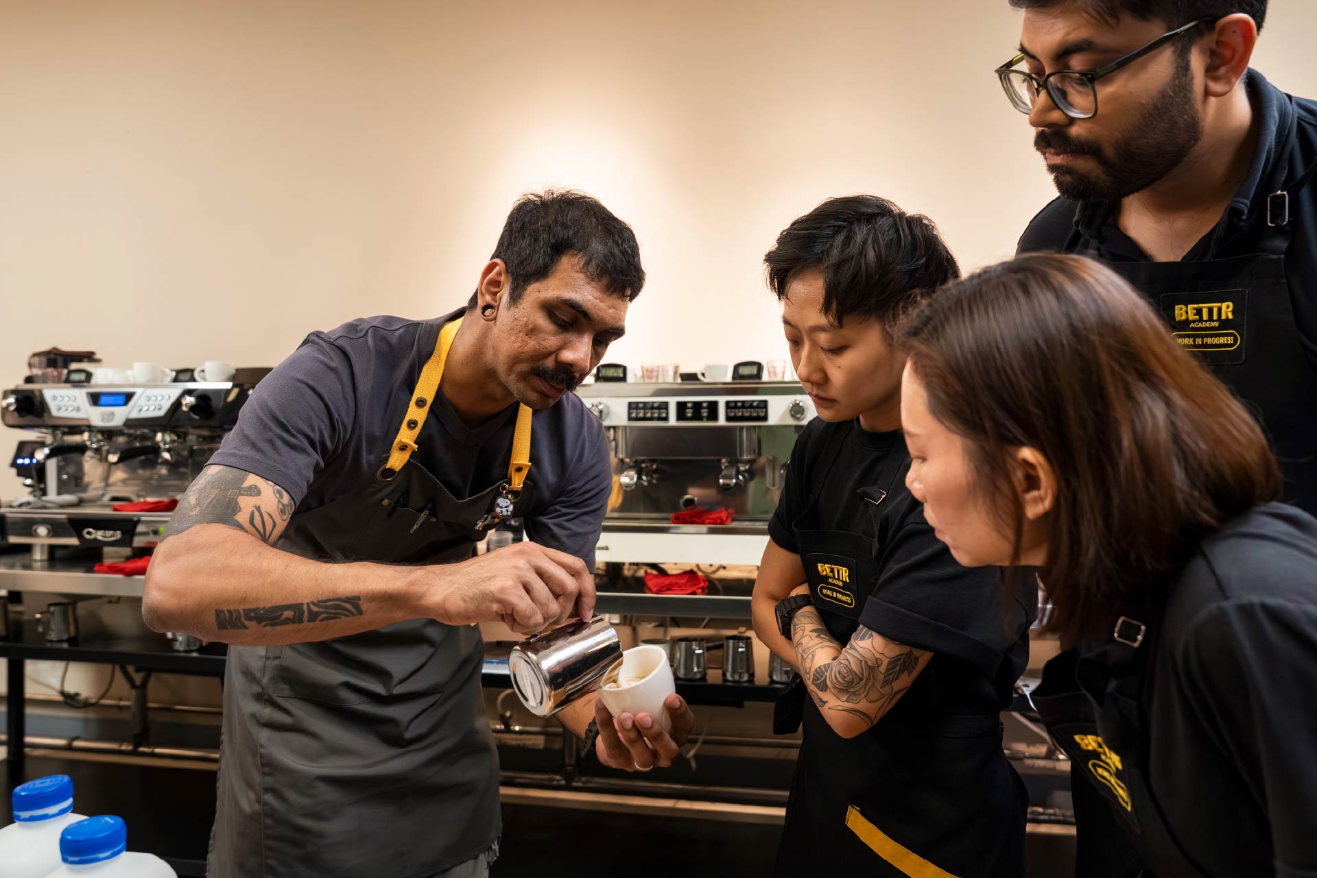 Barista instructor demonstrating latte art pouring milk as three students watch closely in a coffee training class.
