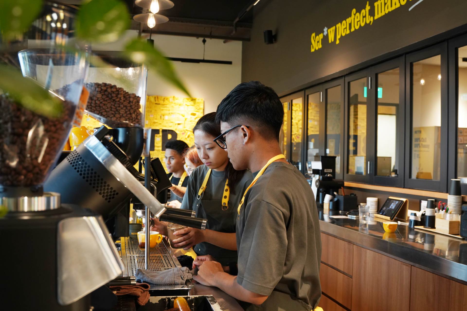 Baristas in gray shirts and yellow aprons preparing coffee drinks behind a cafe counter.
