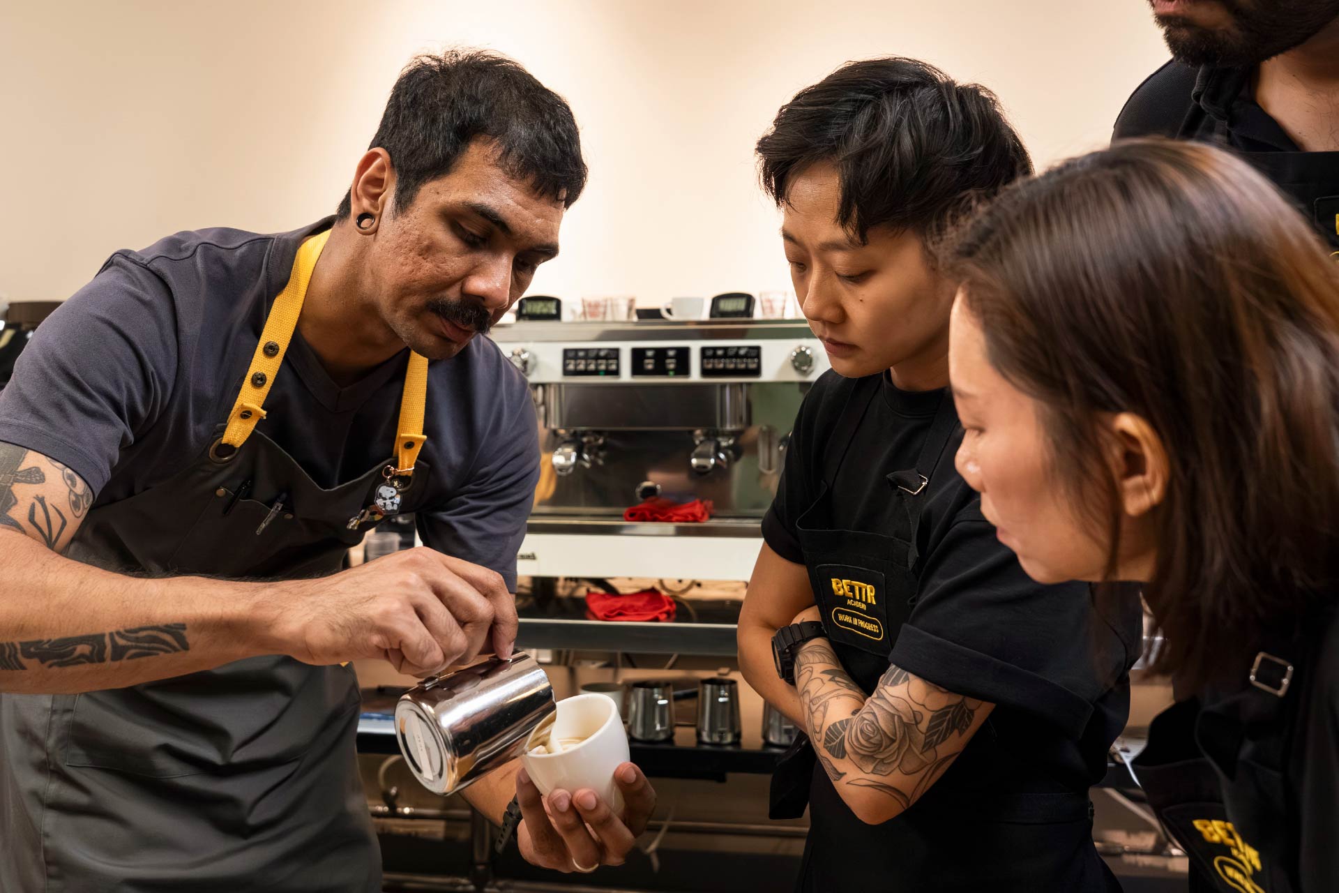 Barista with tattoos demonstrates latte art pouring to attentive trainees at coffee machine.