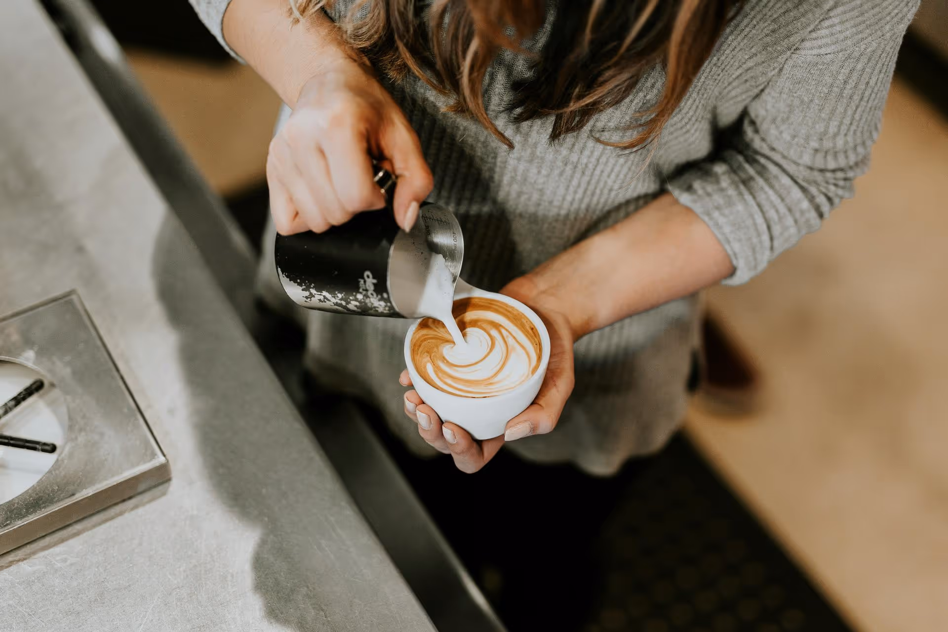 Person pouring steamed milk from a black pitcher into a cup of coffee with latte art.