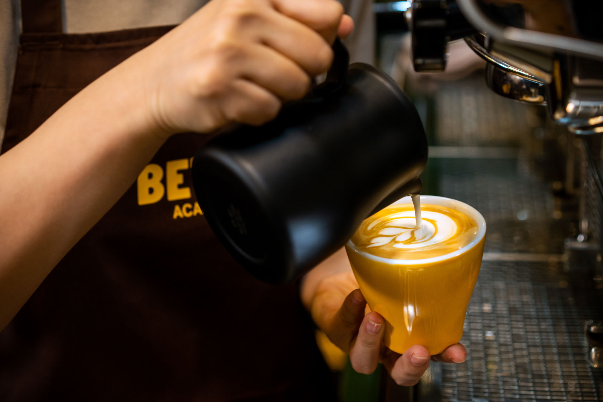 Hands holding spoons, tasting multiple cups of black coffee on a metal table.